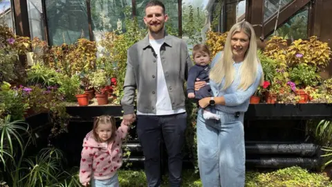 Julie Keegan A family of four inside a greenhouse in a botanical garden with lots of plants in the background. In front of these is a toddler with a pink, furry jacket with love hearts is holding the hand of her father in navy chinos, white t-shirt and grey jacket that is open. A baby boy in a navy outfit is being held by his blonde mother wearing a blue fluffy jacket and jeans.