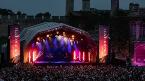 A night-time view of a performance at Lincoln Castle. A large crowd is illuminated by red and pink dazzling neon lights. They are in front of the stage. Two performers are lit by a hazy purple light. 