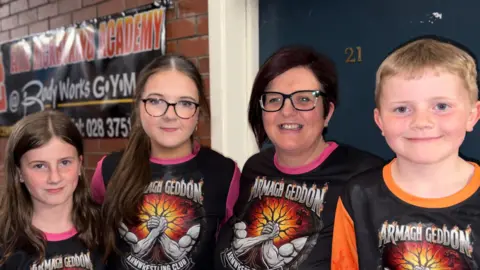 A young girl with brown hair, another young girl with brown hair and glasses, a lady with dark purple hair, glasses and a young boy with blonde hair. 