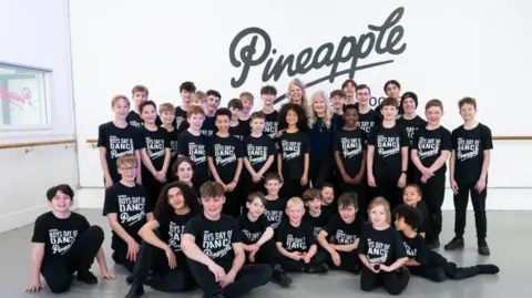 Jayne Jackson 40 male dancers all looking at the camera and wearing black dance trousers and t-shirts with white writing on. Some of the team are standing, whilst others are sitting on the floor all in front of a white wall with a black Pineapple logo.