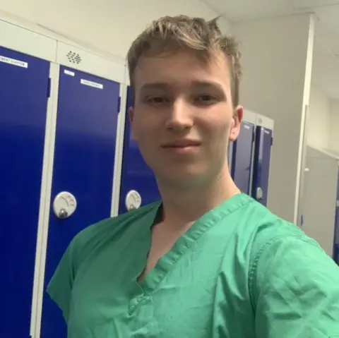 Harrison Sharples A selfie of Harrison dressed in scrubs. The photo is taken in the changing room, the lockers are seen on the background.