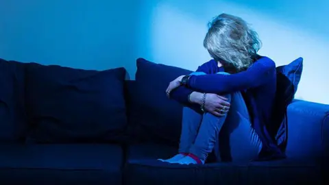 Generic image of a woman sat curled up on a blue sofa with light coloured hair and dress in blue clothing with the room bathed in blue light