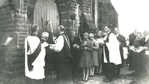 The Bishop of Blackburn at the doors of the new church of St Stephen's Grane after its move in 1927 to officially open it with other church officials and parishioners standing behind him reading a book during the service