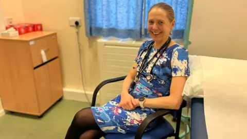 Lucy Gossage Lucy in a blue floral dress, sitting on a chair, with a stethoscope round her neck
