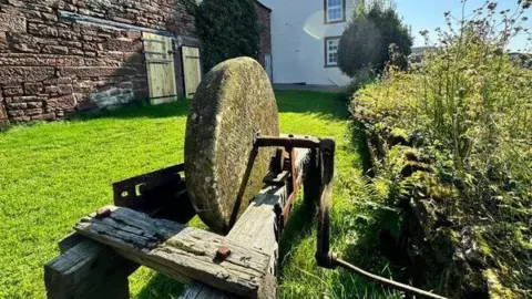 A large sandstone wheel pictured sitting on an old wooden oak frame, in a garden on a sunny day.