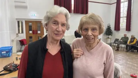 Left Carol to Right Judy two older women standing smiling at the camera.  Carol is wearing a red jumper and black gilet while Judy is wearing a pink jumper.