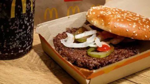 Reuters A McDonald's Quarter Pounder in a box next to a plastic cup filled with Coca Cola. The burger has two buns, one of which is seeded, and it has a beef patty, slivered onions, and gherkins in it.