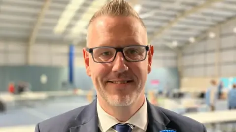 A close-up image of a man with short blonde hair who is wearing dark-framed glasses, a cream-coloured shirt and blue tie and a grey suit. The room behind him is blurred.