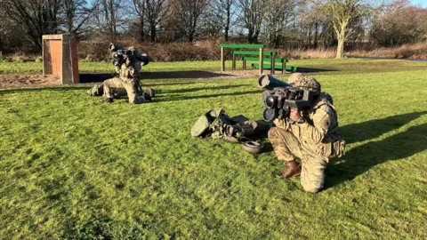 Two soldiers in combat gear outside on grass, on one knee. They are holding a large cylinder on their shoulder, and looking through a sight.