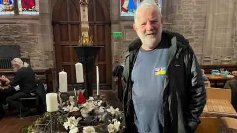 A man with grey T-shirt and black jacket stands in a church hall. Tables and chairs and candles can be seen in the background.