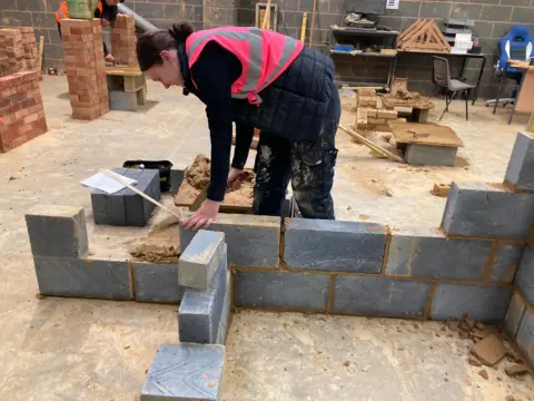 A woman constructing a block wall by applying muck with a trowel 