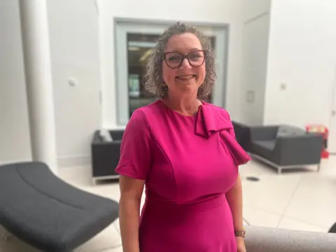 Nadine Peatfield Labour derby city council leader wearing a pink dress and red glasses. She is smiling and stood in a white-floored room.