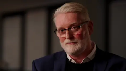 Jim Pearson, a man with grey hair and a grey beard, looking at the camera. He is wearing glasses, a white shirt, a black jumper and a dark blue jacket.