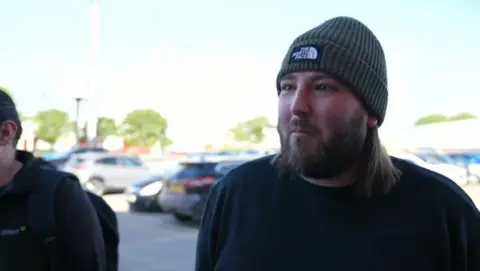 BBC A man with a beard is wearing a hat and a dark top. A car park is behind him.