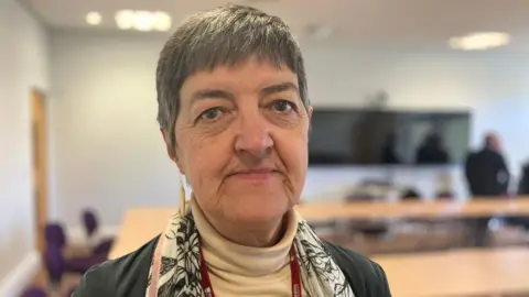 Council leader Lynn Denham in a close-up photograph. She is wearing a beige polo neck and scarf and jacket. She has short hair and is wearing earrings. She is in a council room with people behind her at a table.