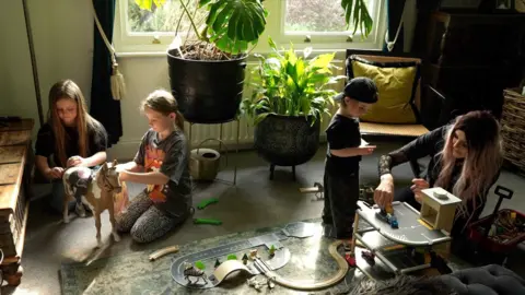 Two girls are sitting playing with a toy horse. A woman and a small boy are playing with a set of cars and a wooden parking garage. There are plants and a window in the background.