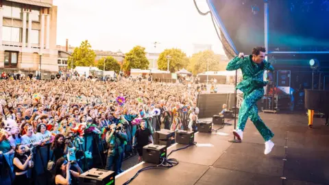 Ania Shrimpton Mika jumping on stage wearing a glittery green suit in front of a huge crowd
