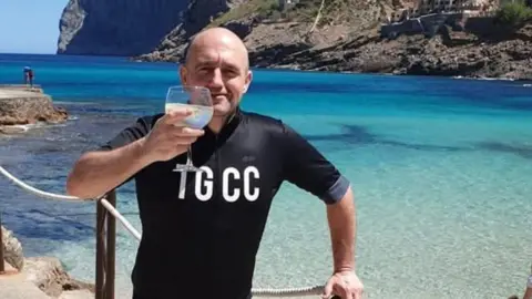 A man standing on a path next to the sea. His left arm is resting on a wooden post and he has a glass in his left hand. He is wearing a black shirt with 'TGCC' written in white capital letters on his chest. 