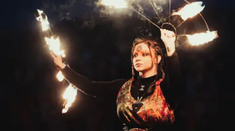 Visit Isle of Man & David Lovelady A woman with her hair in plait, with a sun symbol on her forehead. She holds up two metal semi-circular frames, with fire at the ends of spokes. It is dark and she is lit by the fire around her.