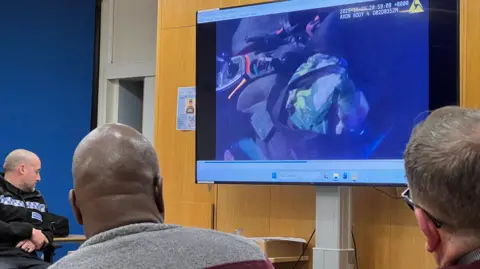 The backs of two people's heads as they watch a large screen showing the inside of a car. A tool can be seen next to the gear stick. Also seen to the left of the two men viewing the footage is a police officer.