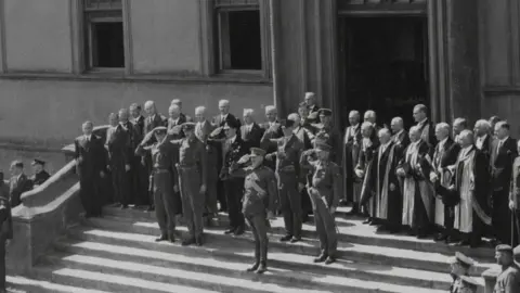 The British Channel Islands WWII Remembrance Campaign Brig Snow is standing in full military uniform on steps alongside other military personnel. They are saluting. Behind them is a large building. The image is black and white. 