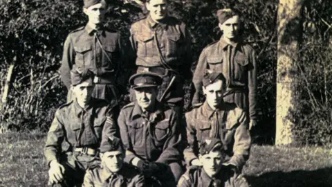British Resistance Museum A blurry black and white photo of the Stratford St Andrew patrol before they disbanded in 1944. It shows eight men in Home Guard uniform, three standing at the back, three sitting on chairs in the middle and two sitting on the grass. Behind them are bushes and a tree trunk. Back row: Albert Cable, Hector Wade, Alexander Dunnett, with Stanley Crane and Arthur Whiting in the front row.