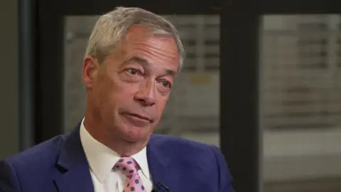 Nigel Farage is wearing a blue suit and patterned pink tie and is seated indoors, framed from the chest up, with frosted glass panels in the background, during the BBC interview.