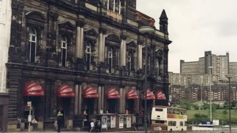 www.picturesheffield.com. To the left of the image; Canada House in the 1980s, with dark, dirty brick work and 7 red canopies over each doorway and window on the front of the building.  Outside there is an empty bus stop, and a bus shelter with passengers waiting, and a cream coloured bus with a brown roof moving away from the bus shelter.  In the back ground, and to the right of the image is a large green space with trees and then three rows of flats with an imposing Park Hill in the background.