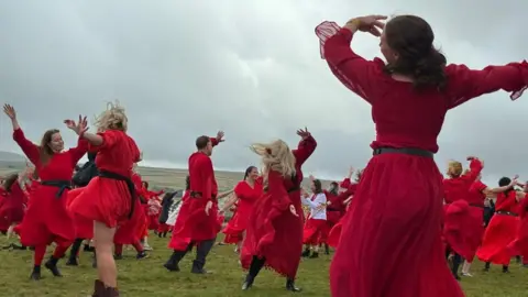 A group of people dressed in red robes dance in a grassy field.