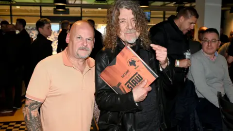 Ed Bagnall Two men, one bald, one with long curly hair and holding a copy of a book about football shirts 