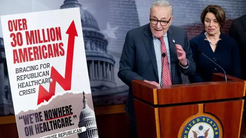 Chuck Schumer behind a lectern with a sign saying that 30 million Americans are bracing for a healthcare crisis. 