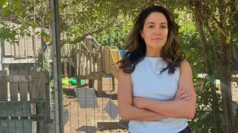 A brown haired woman stands with arms crossed outside a fenced play area at a nursery, with children’s play equipment visible behind