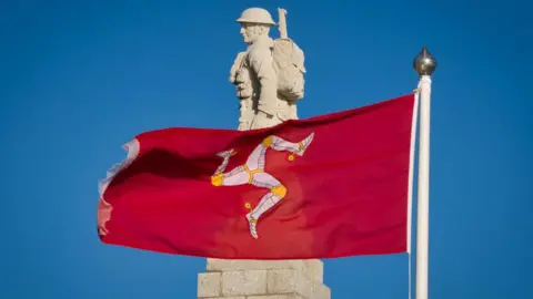 The stone carved World War One soldier that stands on the top of the Douglas war memorial with a red Manx flag flying across it, set against a clear blue sky.