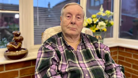 Dolly Carter/BBC Mike Sarre, wearing a navy and purple checked shirt, looking into the camera from his flat in Norwich, with the window behind him