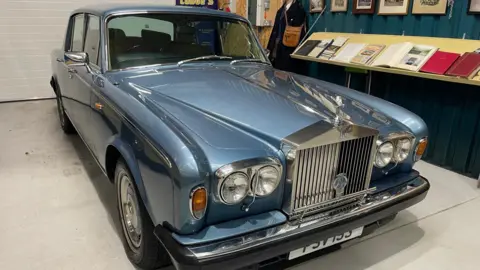 Steve Huntley/BBC The blue Rolls Royce sits on display in the museum. An exhibition to its right with books and pictures can be seen as well as a dummy wearing an old bus driver uniform.