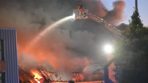 Pacemaker A fire fighter is on a crane in the air and water from a hose is being poured onto a fire. Metal coverings are lying about.