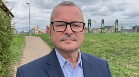 Sam Read/BBC A head and shoulder shot of Lee Barron. He has black-framed glasses and is wearing a mid blue jacket over a light blue open necked shirt. Behind him is grass, a path and a children's play area behind a fence. 