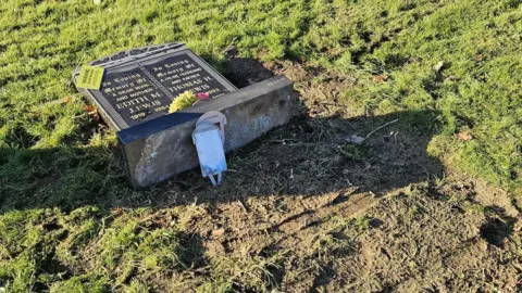 Family Handout A grave stone knocked over.  On the black stone is two names in gold lettering. There is a yellow warning sign on the top left corner and some flowers at the bottom. Around the stone is grass and muddy soil with tracks in it. 