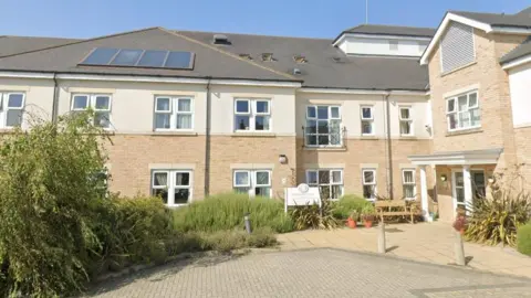 The front of Handford House care home building. Bushes are outside with a doorway on right hand side of image. Building is two stories tall with numerous windows. A sign is in the centre of the image. 