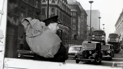 PA Media An old black and white photo of postman Rocky carrying his parcels in a sack on his back to hand-deliver them. Cars and iconic London red buses can be seen in the background