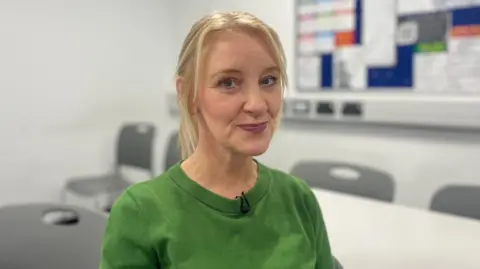 Head and shoulders picture of Liz Baxter who is wearing a green top. Grey chairs and a white table can be seen in the background.