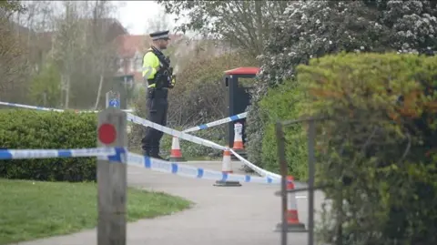 Jamie Niblock/BBC Blue and white police tape is attached to cones and posts i a park. A police officer in uniform stands in the background.