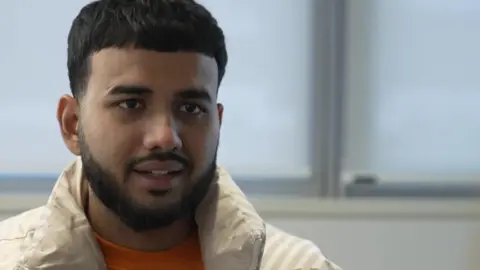 Sojibe Hossein, a young man with dark hair and a beard in a cream puffer jacket and orange t-shirt. He is sitting in an office