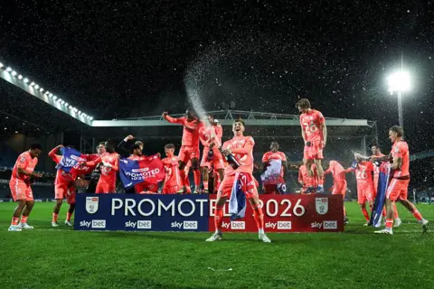 PA Media Coventry City players on the pitch post-match. Many of them are behind a board reading "Promoted 2026". Some have flags others bottles of Champagne