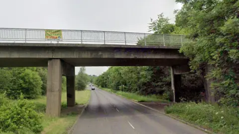 A concrete bridge with a small yellow banner hanging off the railing overlooks a road.