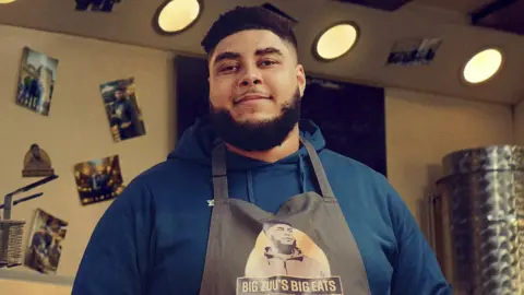 BBC/FBO Big Zuu from his BBC series Big Zuu's Big Eats. Big Zuu is a 28-year-old black man with short afro hair, brown eyes and a dark beard. He wears a grey apron with 'Big Zuu's Big Eats' printed on it over a blue hoodie. He's pictured inside a kitchen which has photos printed on the walls.