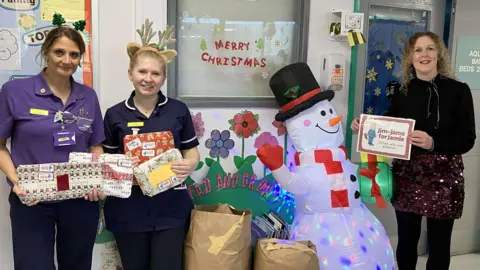 Three woman standing in a hospital corridor with large bags between them containing gift-wrapped pyjamas