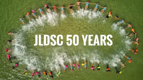 JLDSC An aerial photo looking down into the sea. The water has a green tinge. Multiple members of the Jersey Long Distance Swimming Club are floating in the water in a long circle shape, all holding hands. In the centre of the circle the text 'JLDSC 50 YEARS' has been edited onto the image.