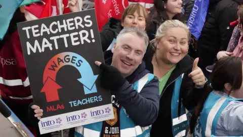 PA Media A man holding a sign that's reads "teachers matter!" and has an arrow with the word "wages" on it pointing from "MLAs" to "teachers" The Sign also has INTO branding. Beside him is a woman smiling and giving a thumbs up to the camera. They are standing in a crowd of people.