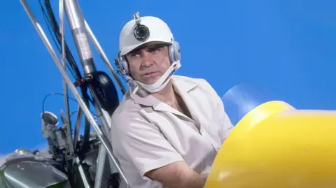 A photo taken from the James Bond film You Only Live Twice. It features actor Sean Connery in a yellow autogyro. He is wearing a short sleeved shirt and is wearing a helmet with a camera attached to it.